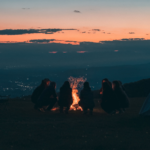 Eine Gruppe Menschen sitzt in der Abenddämmerung um ein Lagerfeuer und erzählt sich Geschichten
